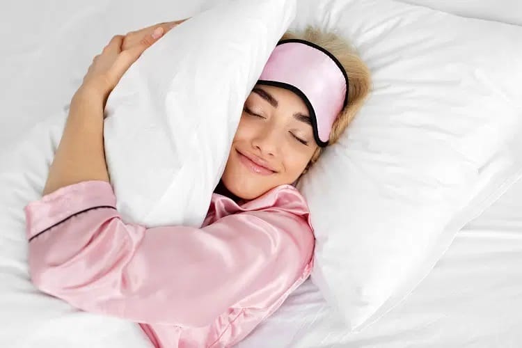 Femme en pyjama rose avec masque de sommeil sur le front et yeux fermés serrant une couette dans ses bras
