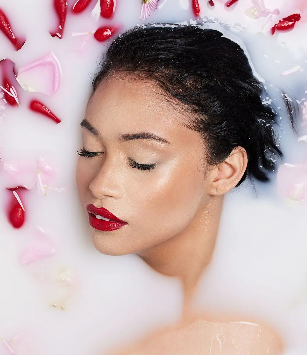 Gros plan sur le visage d’une femme au rouge à lèvre rouge allongée dans un bain de lait avec pétales de rose rouge autour d’elle